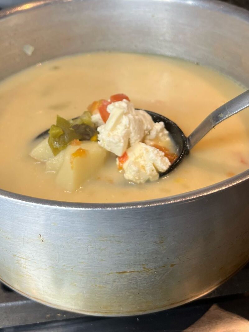 A close-up of a metal pot containing creamy soup with chunks of chicken, potatoes, and colorful vegetables, being served with a ladle.